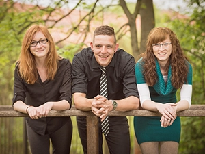 Drei lächelnde Geschwister, zwei Frauen und ein Mann, lehnen entspannt an einem Holzgeländer im Freien. Ein lebendiges Gruppenporträt, ideal für Geschwister-Fotoshootings und Familienreportagen in Erfurt von Michael Schalansky Fotografie.
